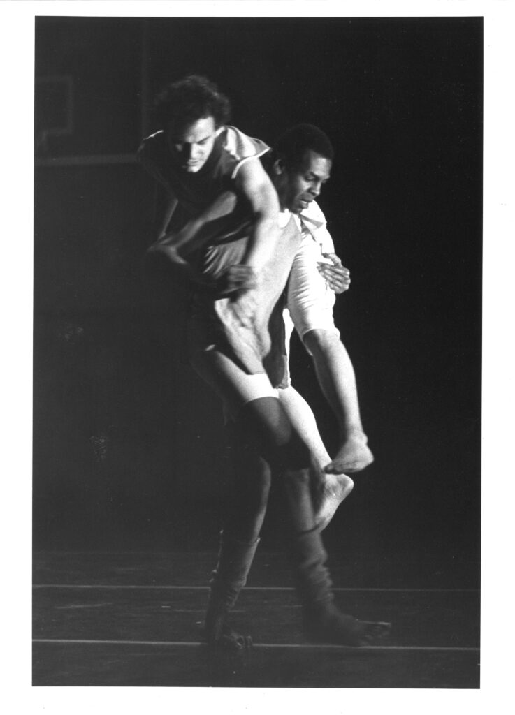 In a black and white archival photo, Gus walks to stage left, eyes focused down as he carries Toby over his shoulders.