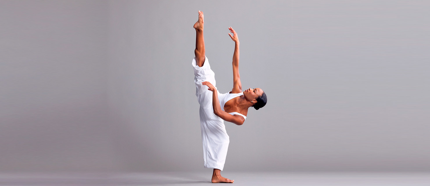 Alexandria Best extends her leg to 180 degrees to the side, standing leg in plié. Her torso leans gently away from her raised leg, arms softly extending as she twists to face it, face upturned. She is barefoot and wears white, the leg of her pants cuffed to her knee on her extended leg.