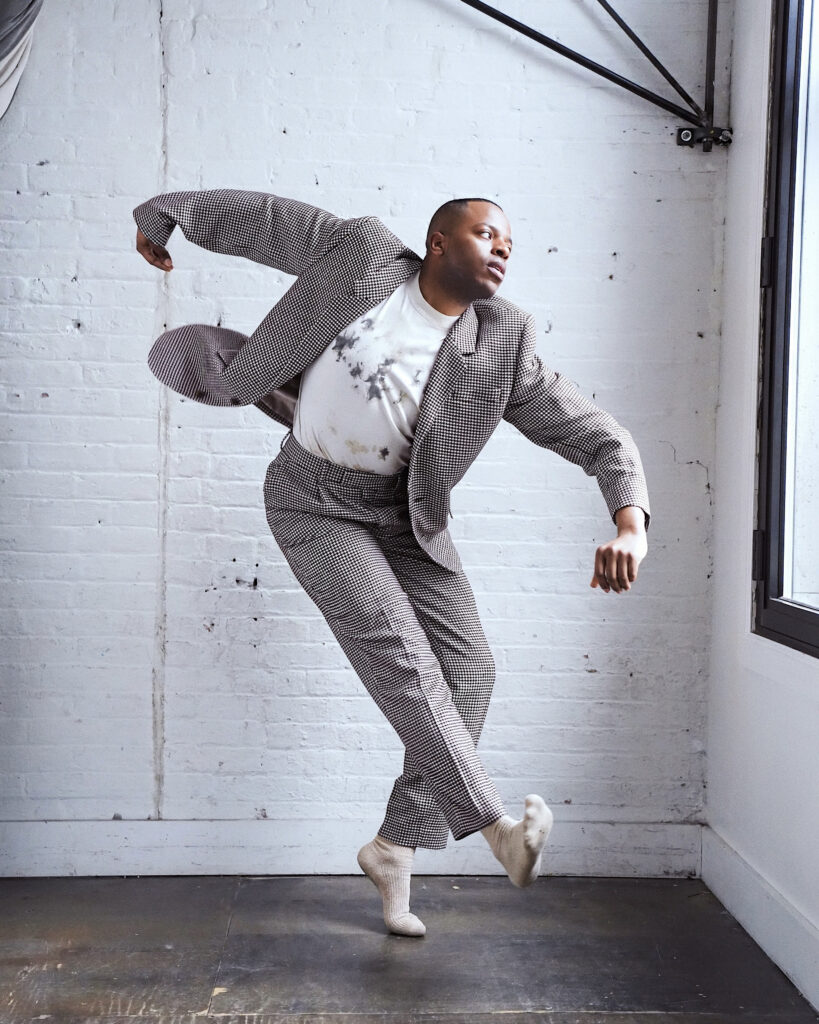 a male dancer wearing a brown suit and posing in a white room