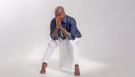 Hope Boykin, a petite, curvy Black woman with a shaved head, gives the camera a mischievous, close-mouthed smile. Her palms are pressed together with her fingertips resting against her lips; her elbows are propped on her knees as she leans forward where she sits, arches popping against the floor. She wars white trousers and a denim button-down with the sleeves rolled to her elbows.