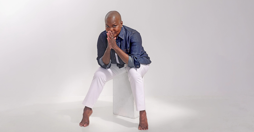 Hope Boykin, a petite, curvy Black woman with a shaved head, gives the camera a mischievous, close-mouthed smile. Her palms are pressed together with her fingertips resting against her lips; her elbows are propped on her knees as she leans forward where she sits, arches popping against the floor. She wars white trousers and a denim button-down with the sleeves rolled to her elbows.