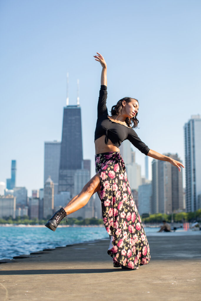 a female dancer wearing a long skirt posing with one leg extended front with a city skyline behind her