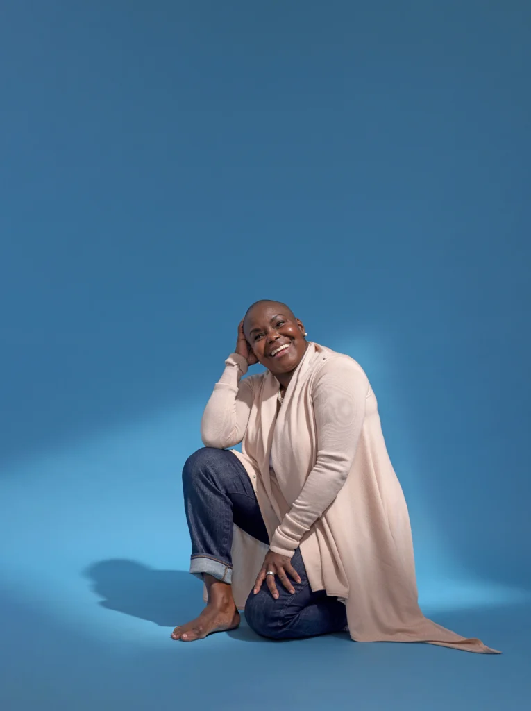 Hope Boykin, a petite, curvy Black woman, laughs as she looks to a corner. She kneels facing the side against a blue backdrop. One palm rests against the side of her shaved head, that elbow propped on her knee. She wears a draping off-white cardigan and cuffed blue jeans.