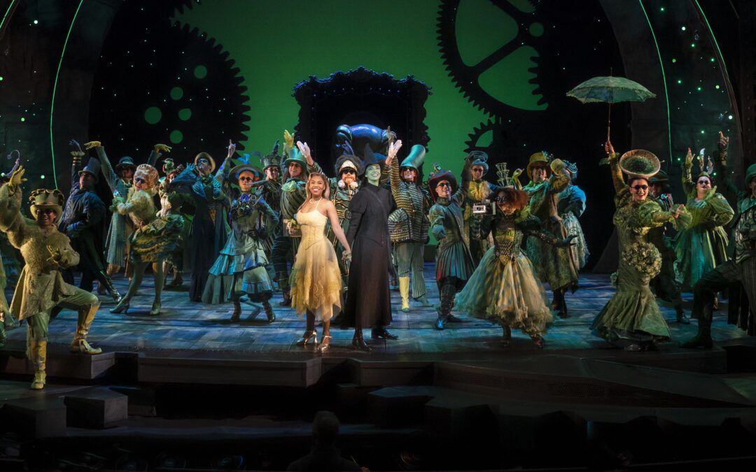 a group of performers on stage, one in a yellow dress, the other in black with green skin, the ensemble behind them