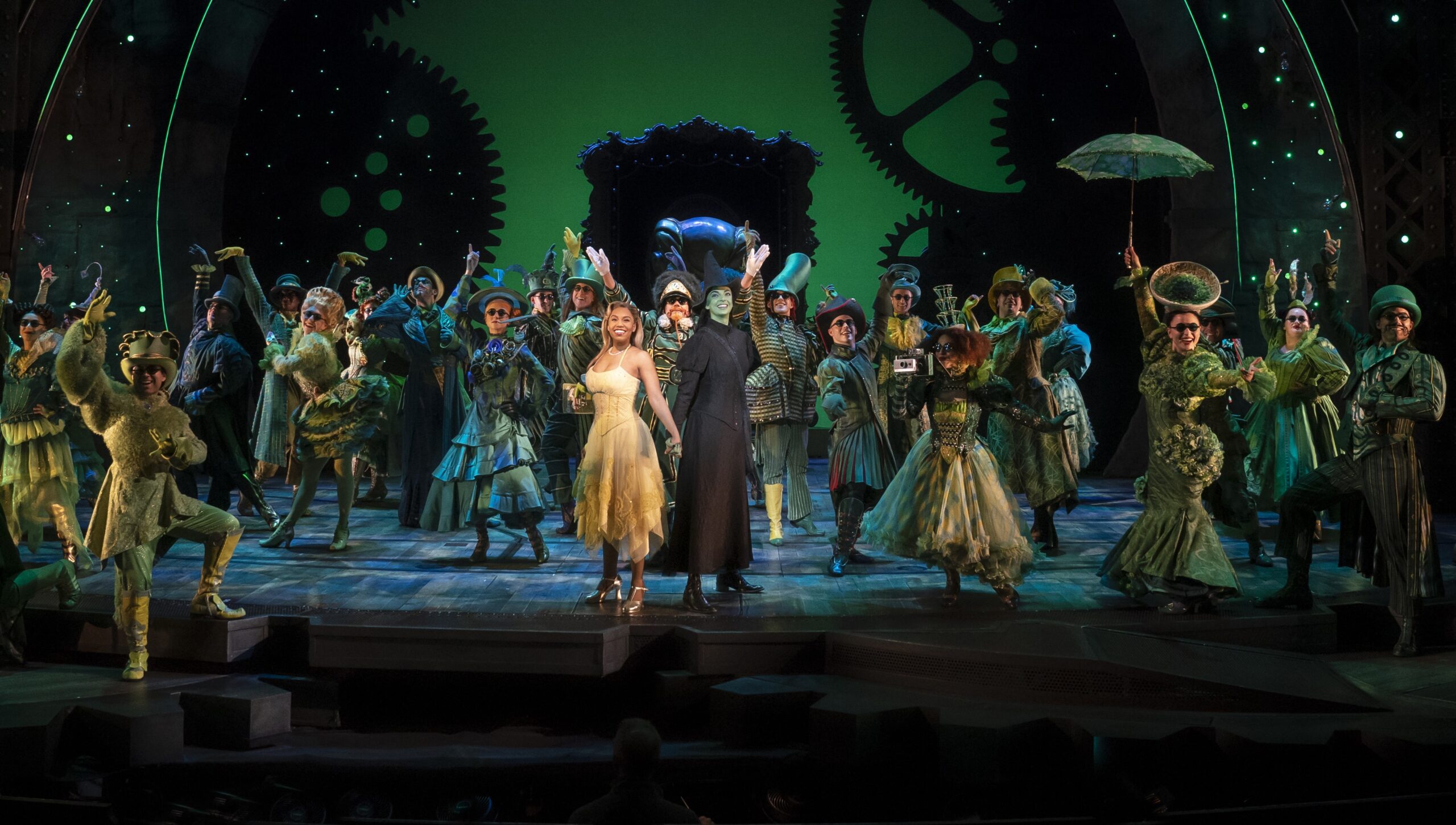a group of performers on stage, one in a yellow dress, the other in black with green skin, the ensemble behind them