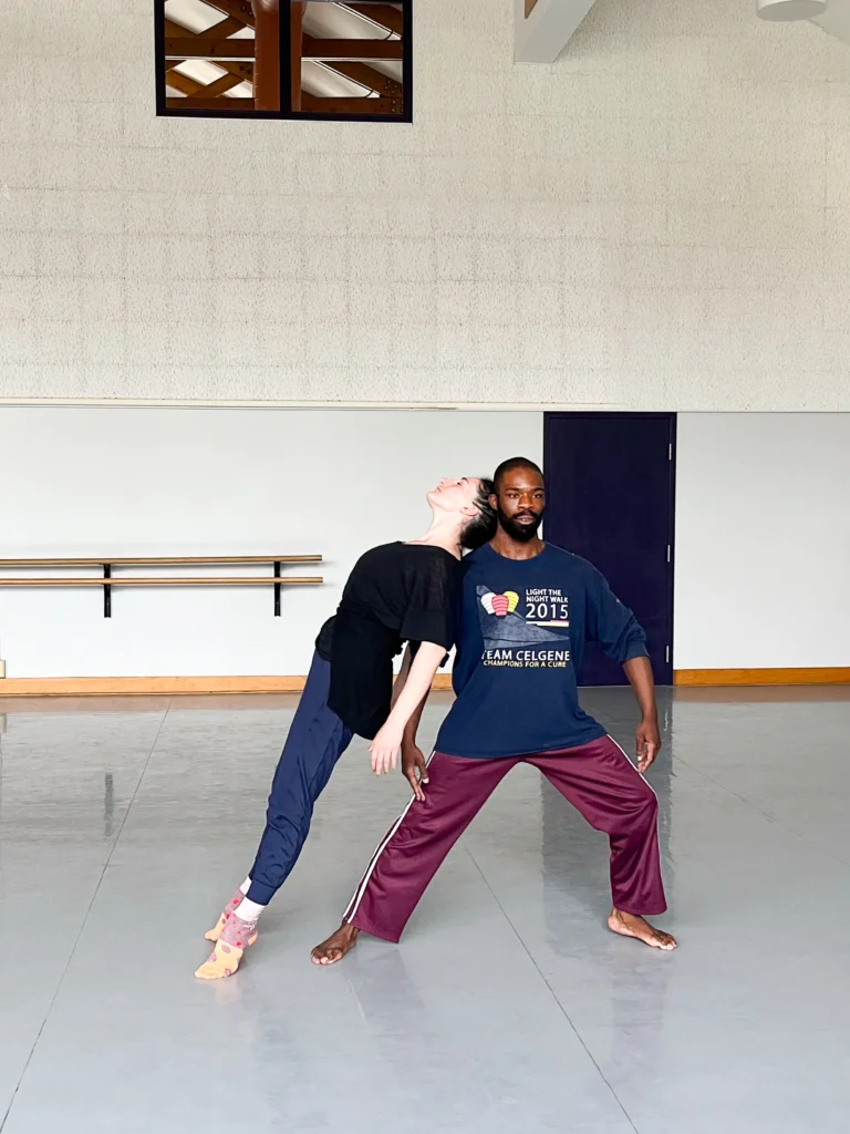 A man in a dark blue t-shirt and burgundy sweatpants lunges to the side while a woman in dark blue pants and black top arches back leaning on the side of the man.