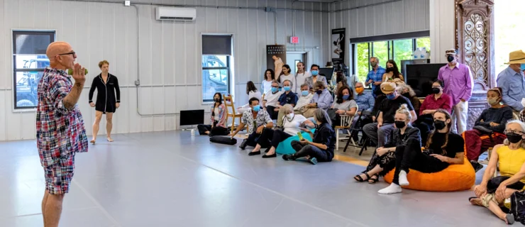 Stephen Petronio stands barefoot in a spacious studio as he addresses a small audience seated in chairs and on the floor. A dancer in a black costume reminiscent of a silk pajama set begins to walk from the side of the space.