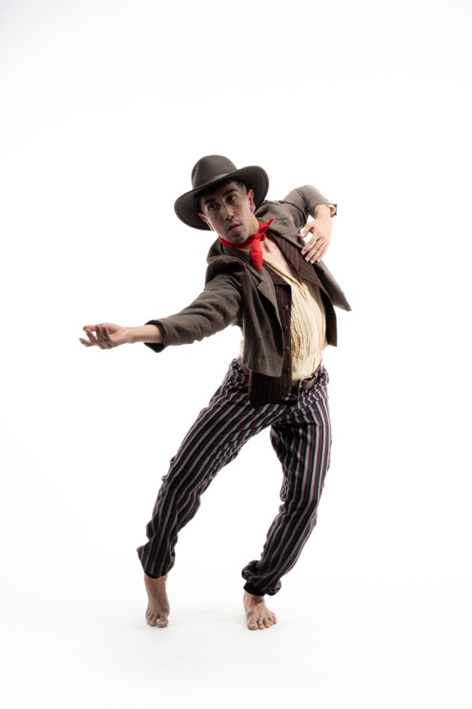 A dancer wearing a cowboy hat and clothes reminiscent of the Old West, except for his bare feet, poses against a white backdrop. His knees bend, one foot moving to forced arch as he twists his torso in opposition. One hand is extended, palm up, the other resting against his chest.