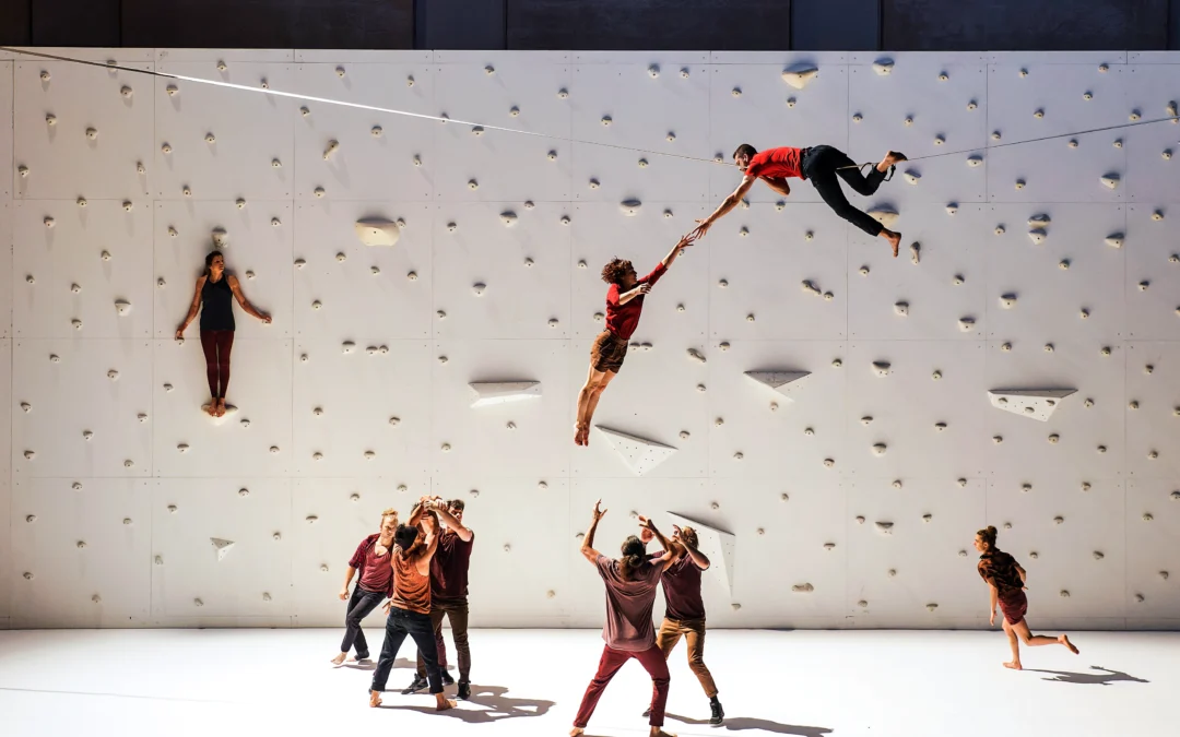 High above a white stage, a dancer lies on his stomach on a tightrope, another dancer swinging down from his hand. Two pairs of dancers stand facing each other below; one set has just flung their compatriot in the air, while the other braces to catch a falling body. Two dancers run in loops at the edges. Another balances on a narrow ledge 10 feet above the stage, back pressed to a rock climbing wall upstage.