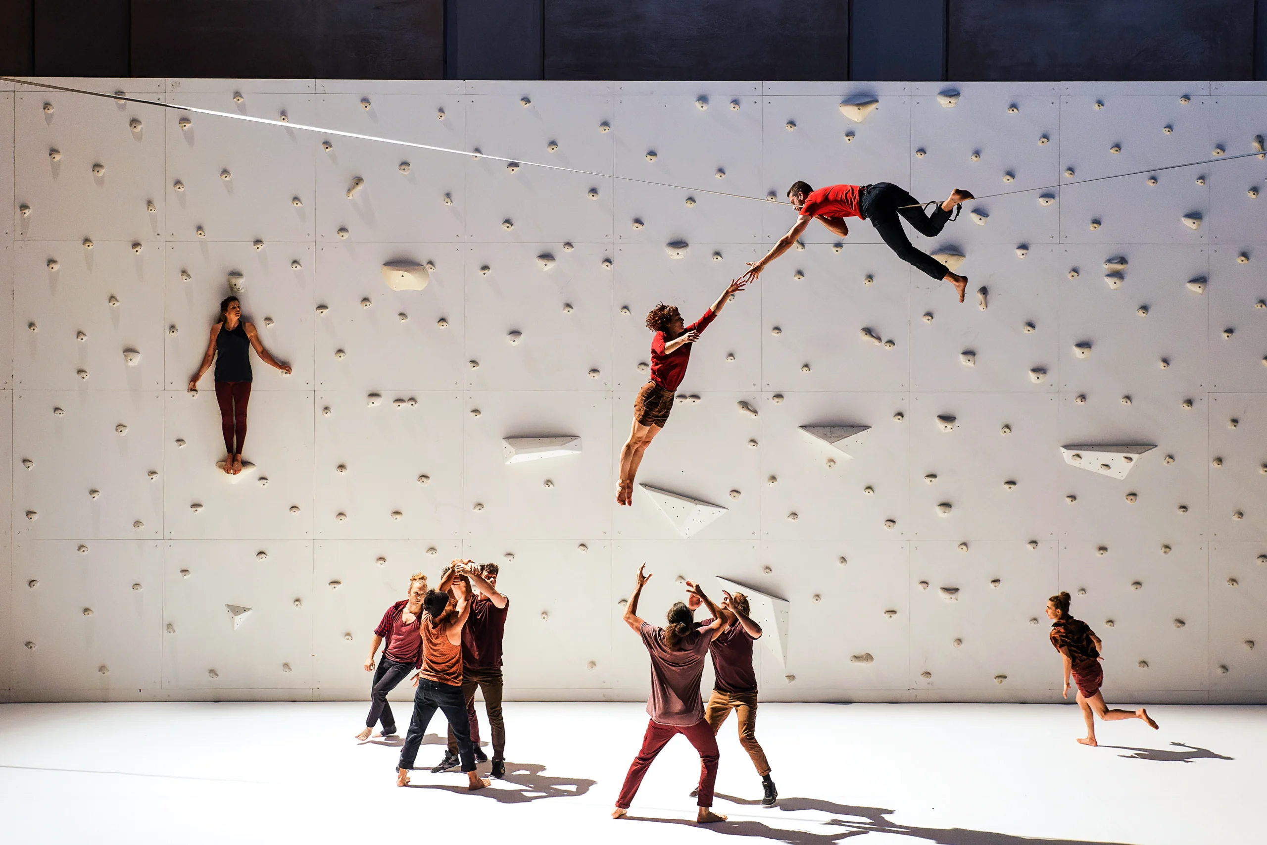 High above a white stage, a dancer lies on his stomach on a tightrope, another dancer swinging down from his hand. Two pairs of dancers stand facing each other below; one set has just flung their compatriot in the air, while the other braces to catch a falling body. Two dancers run in loops at the edges. Another balances on a narrow ledge 10 feet above the stage, back pressed to a rock climbing wall upstage.