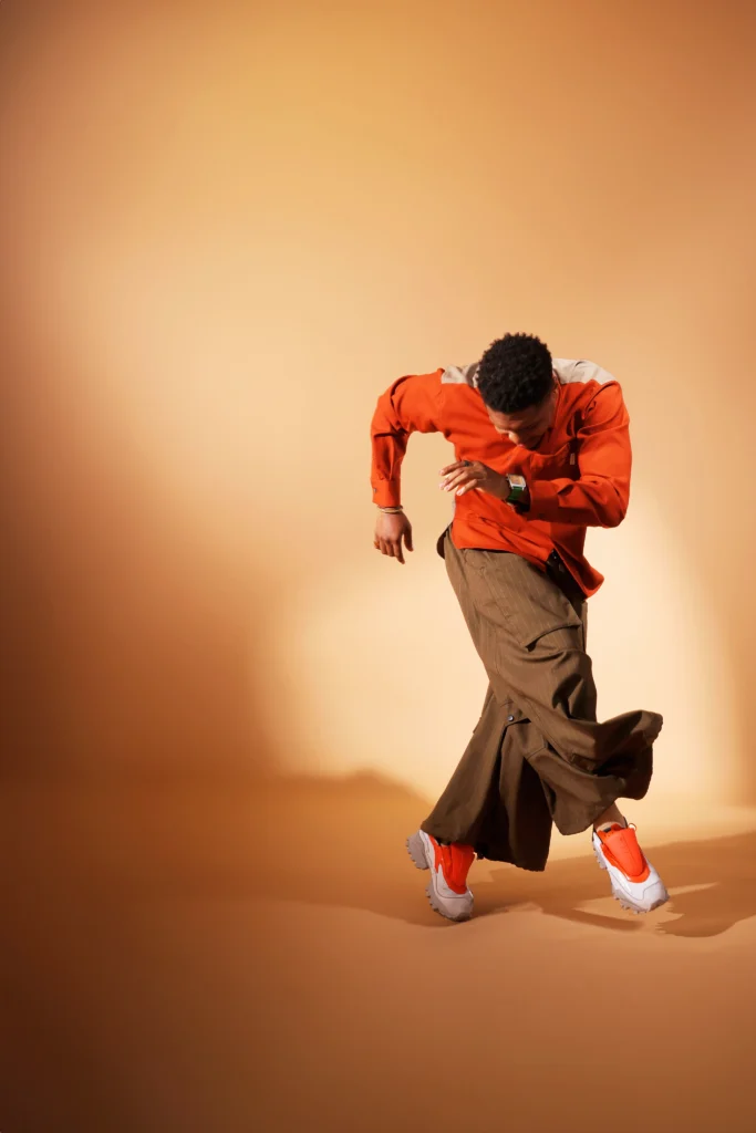 Ephraim Sykes, an athletic Black man in his late 30s, poses against a muted orange backdrop. His head is bowed toward his feet as he moves through a crossed fourth position on relevé, arms angularly working in opposition. He wears a long sleeved shirt that is orange in the front and beige in the back, billowing brown pants, and sneakers with orange laces.
