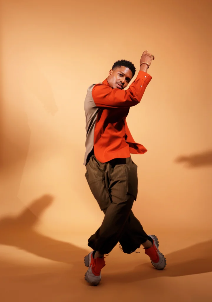 Ephraim Sykes, an athletic Black man in his late 30s, poses against a muted orange backdrop. His legs cross as he rises on forced arch, upper body twisting in opposition so his right arm crosses his torso just below his face. He wears a long sleeved shirt that is orange in the front and beige in theback, billowing brown pants, and sneakers with orange laces.