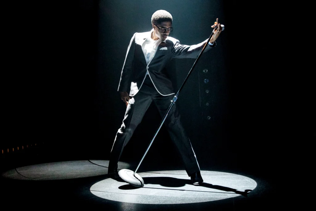 Ephraim Sykes takes a wide stance in a spotlight, catching a microphone stand as it tips over with one outstretched arm. His gaze is downcast, shoulders hitched as he twists toward the mic. He wears a dark suit with white piping and retro-feeling rimmed eyeglasses.