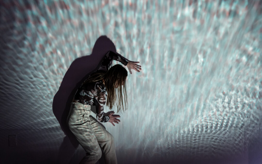 A dancer stands in profile against a wall, slightly stooped over as they press their palms against it, head turned toward it. A projection refracts from and over the dancer, who wears light colored clothing.