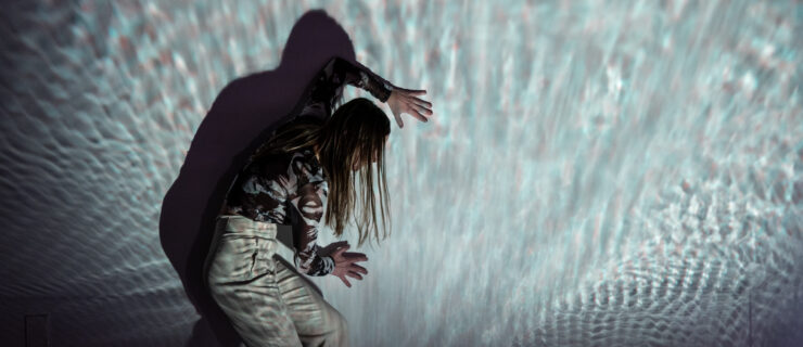 A dancer stands in profile against a wall, slightly stooped over as they press their palms against it, head turned toward it. A projection refracts from and over the dancer, who wears light colored clothing.