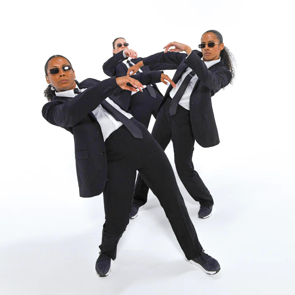 Three dancers in black suits, white shirts, and dark sunglasses lean back in opposite directions from a straight line. It is reminiscent of the bullet-dodging effects in the movie The Matrix.