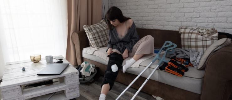 Young woman after an ACL surgery, sitting on the couch adjusting her knee orthosis.
