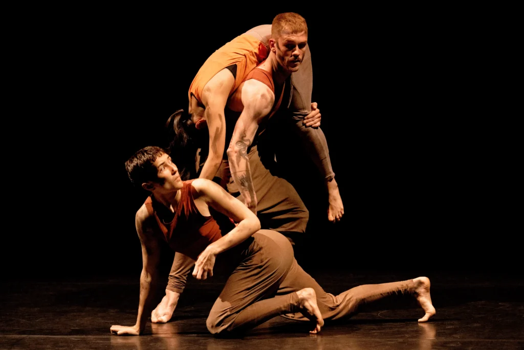 On a dark, sepia-lit stage, one dancer carries another over his shoulder, while a third crawls downstage. The one on the floor pauses, a hand pulled back from the floor as they look over their shoulder, back the way they came.