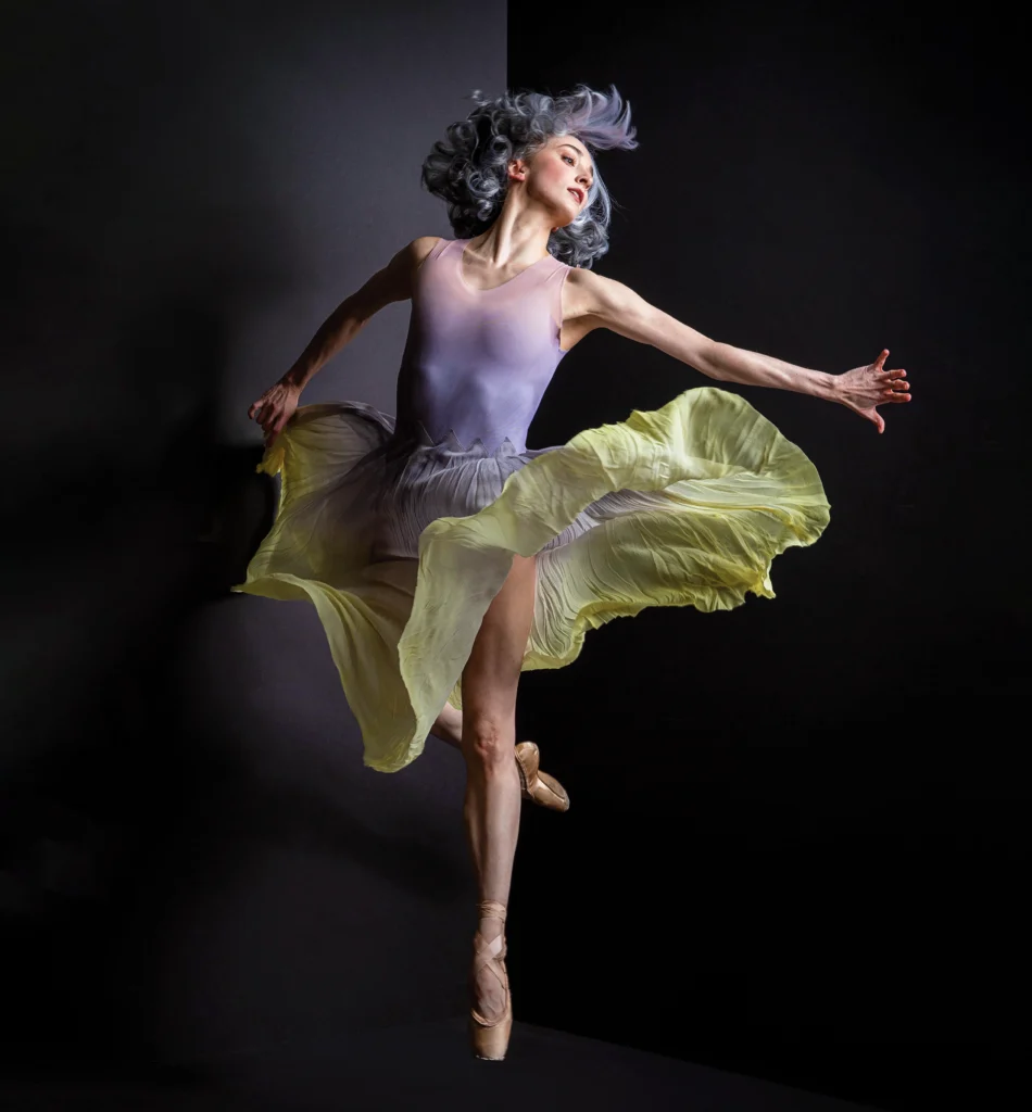 A female dancer turns against a dark backdrop on pointe, raised foot in retiré back, a diaphanous green skirt flaring around her hips and thighs. Her arms reach behind her as she looks over one shoulder, silver hair flying.
