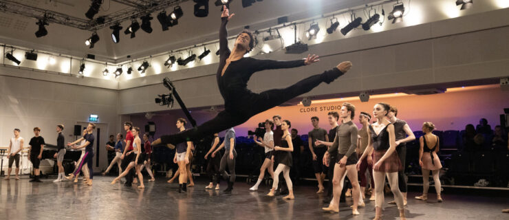 Marcelino Sambé does a flying saut de chat in company class with The Royal Ballet. He smiles jubilantly and wears all black rehearsal wear.