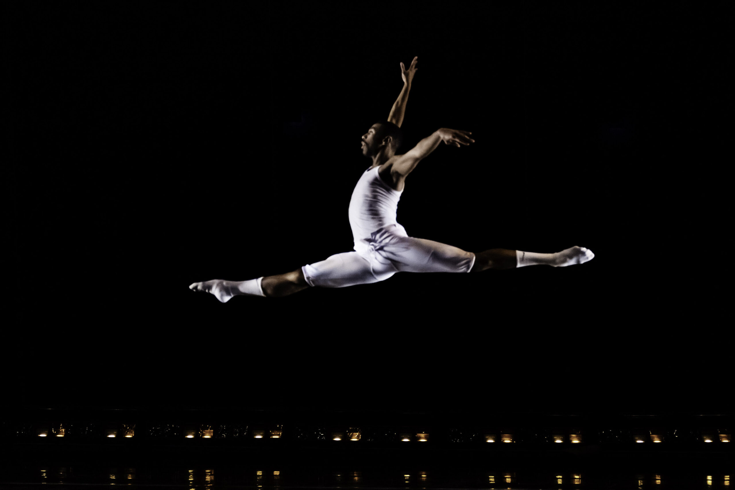 a male dancer wearing white performing a grand jete on stage