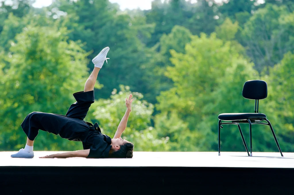 Patricia Delgado dances on an open-air stage backed by lush greenery. She is on her back in a hinge, supported by her shoulders and one foot planted on the ground. Her upstage leg and arm point up to the sky. She wears a black, short-sleeved jumpsuit and white sneakers.