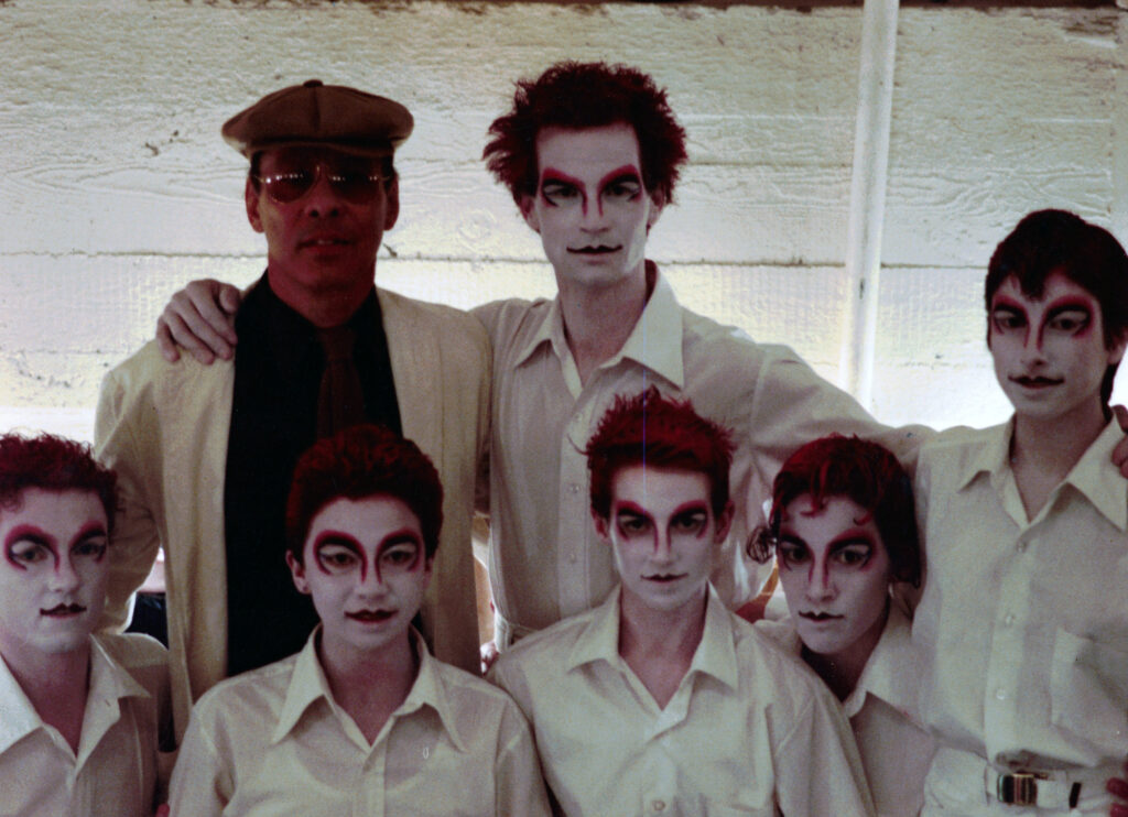 A portrait of seven artists in two rows. Perez, at top left, wears a beret and sunglasses; the others are in white button downs, their faces painted stark white with red accents, their hair streaked with red.