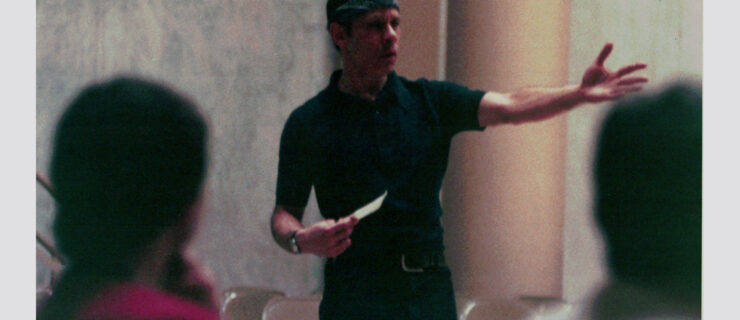 Perez—wearing a dark shirt and pants and a bandanna around his forehead—is pictured mid-speech, gesturing emphatically with his left arm and holding a notecard in his right hand.