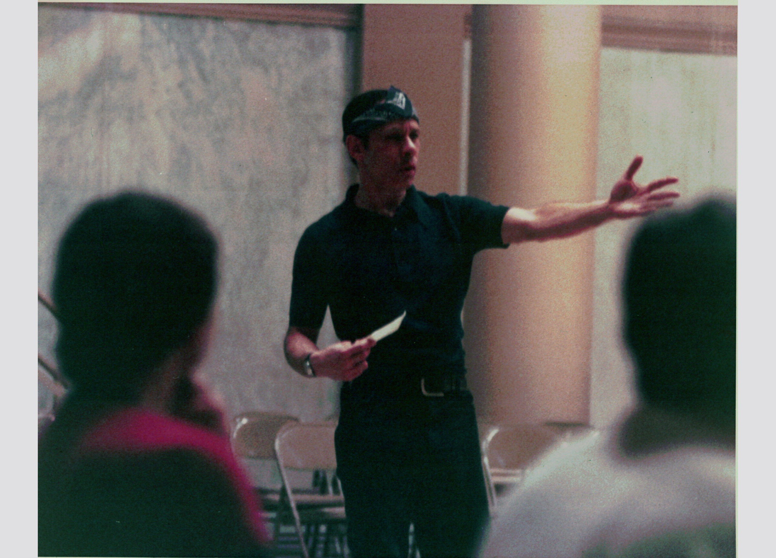 Perez—wearing a dark shirt and pants and a bandanna around his forehead—is pictured mid-speech, gesturing emphatically with his left arm and holding a notecard in his right hand.