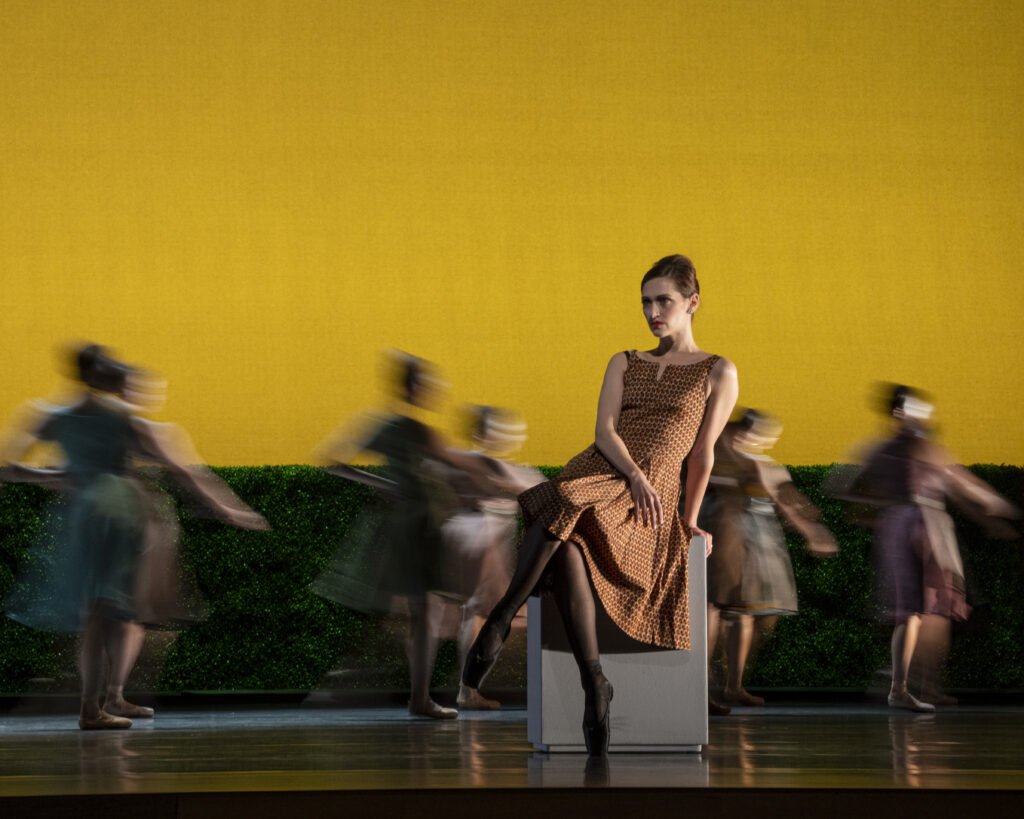 a female dancer wearing an orange dress siting on a block, dancers move in a blur behind her