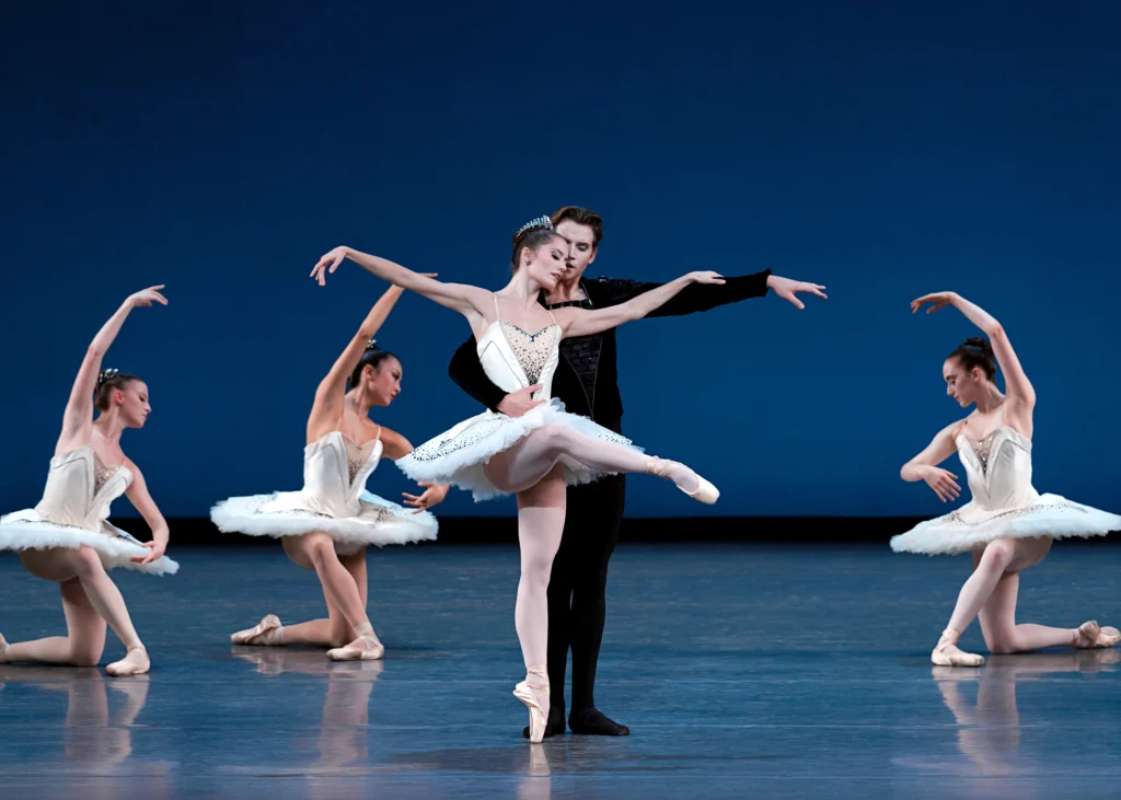 Unity Phelan balances in attitude front on pointe, gaze downcast, one hand resting on her partner's extended arm. Alec Knight stands behind her, helping her balance with an arm around her waist. Upstage around them, dancers in white tutus kneel facing toward the center.