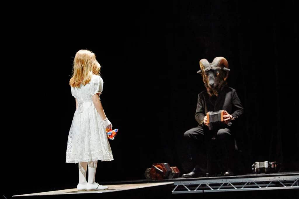 Un jeune enfant blond vêtu d’une robe blanche se tient face au fond de la scène, où une silhouette vêtue d’un costume sombre et portant un masque de bélier inquiétant couvrant tout son visage est assise et joue de l’accordéon.