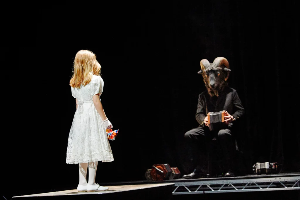 A young blonde child in a white dress stands facing upstage, where a figure in a dark suit and a foreboding ram mask covering their entire face sits playing a concertina.