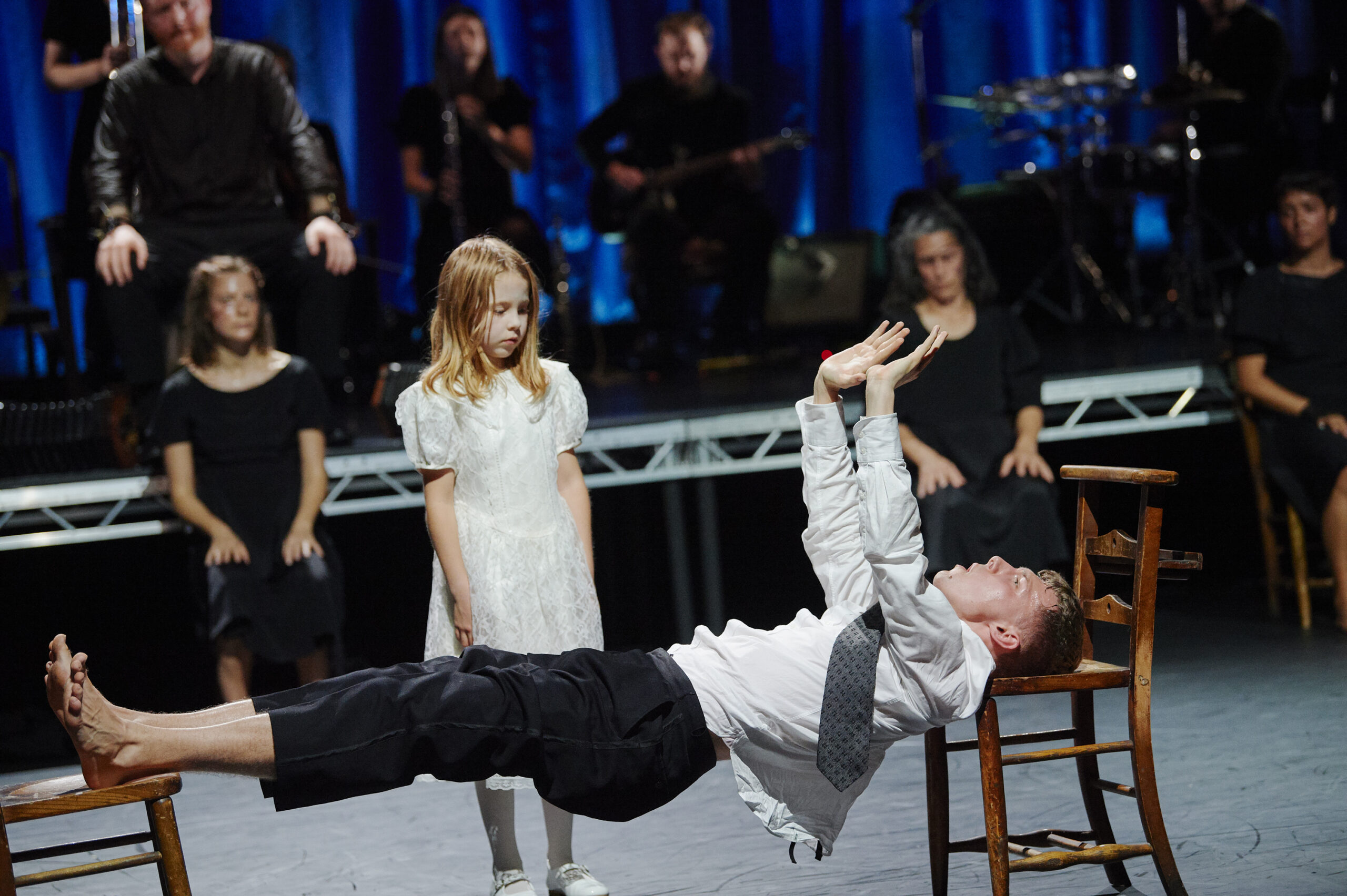 A blonde child in a white dress watches as a male dancer is suspended horizontally between two chairs. He rests his heels on the seat of one and his head on the other, holding his body horizontal to the floor as his palms press into the air above his shoulders. Performers in black sit upstage; others with instruments are on a raised platform behind.