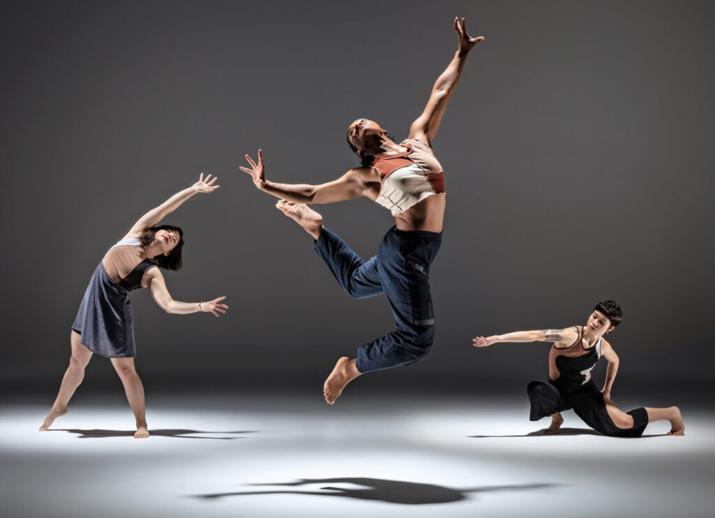 Clarissa Rivera Dyas, a young Black woman, jumps. Her head is thrown back as her arms push back the air around her. Her legs bend beneath and behind her. Two dancers upstage and to either side of her lean in her direction, one standing, the other lunging to one knee.