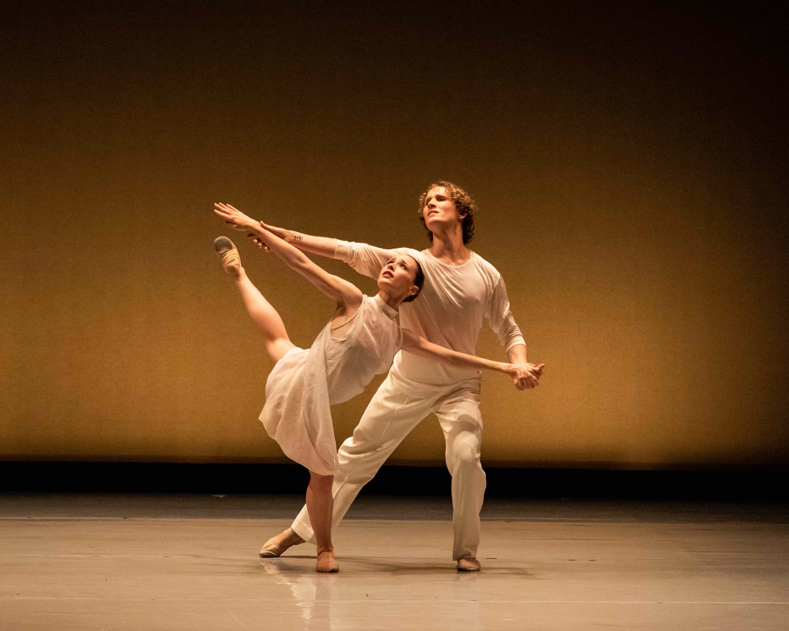 A female dancer moves through a back attitude in parallel, upper body twisting toward her working leg. Her outstretched arms are held by a partner who lunges beside and behind her, gazing in the same upward direction as her.