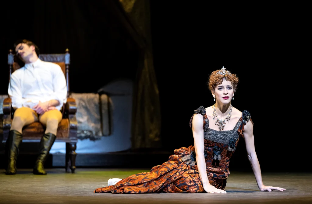 Mayara Magri wears an opulent dress, jewelry, and tiara that are evocative of the turn of the 20th century. She has fallen to the floor and looks out at the audience with a shell-shocked, bitter expression as she pushes herself upright. Upstage, a male dancer sits in a chair, head lolling.