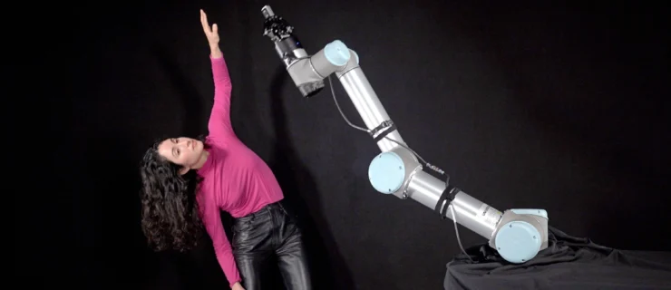 Catie Cuan dances against a dark backdrop, leaning to one side as she raises an arm overhead. In the foreground, a robotic arm imitates the motion as Cuan watches.