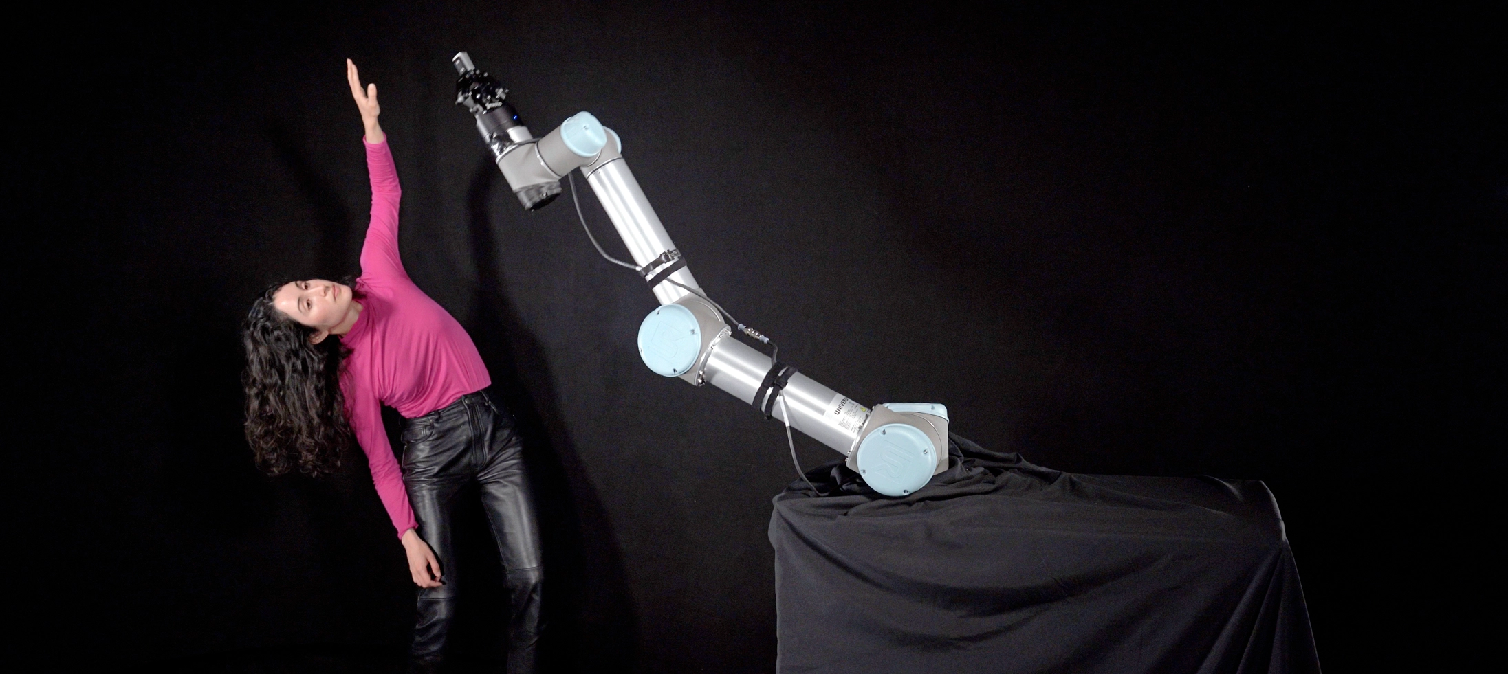 Catie Cuan dances against a dark backdrop, leaning to one side as she raises an arm overhead. In the foreground, a robotic arm imitates the motion as Cuan watches.