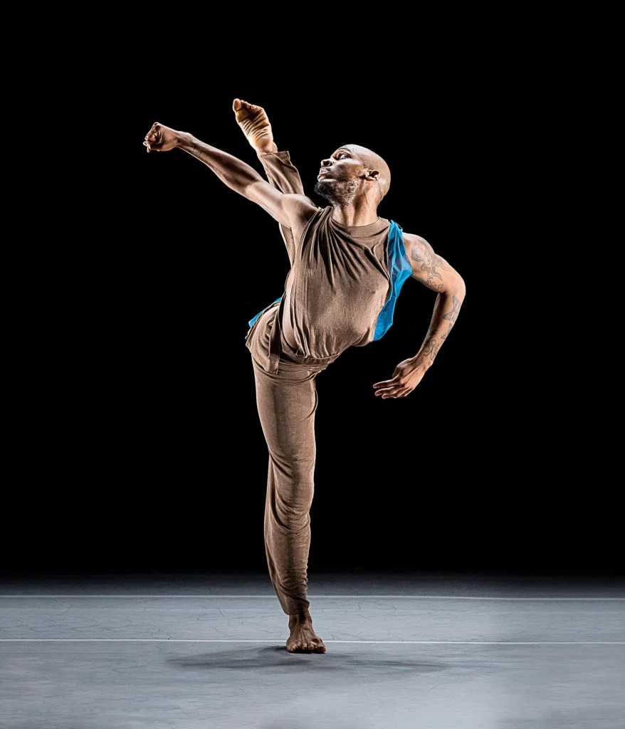 Donovan Reed swings one leg in a parallel attitude behind them. Their opposite arm swings to one side, hand in a fist, as they twist to look over their shoulder toward their back leg. They are barefoot and wear brown pants and a tank top with a strip of flowing blue material. The sleeveless shirt reveals tattoos on their left arm.