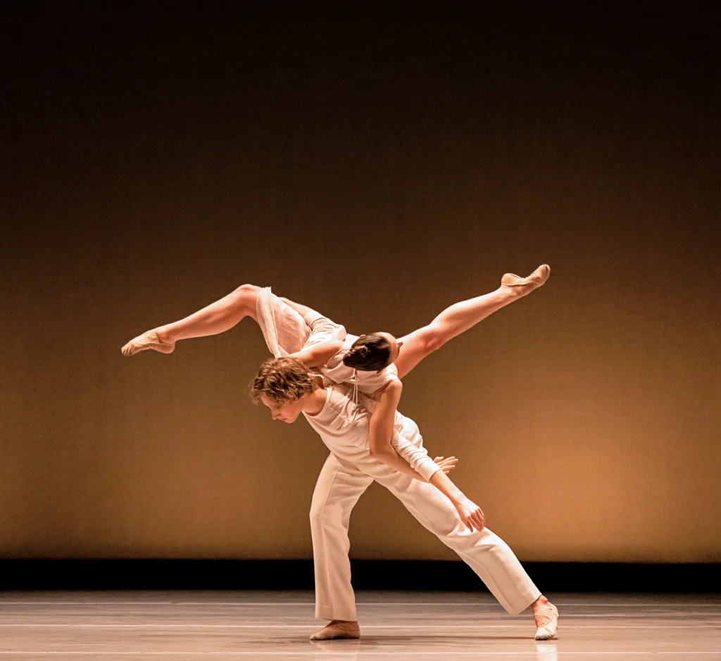 Two dancers are shown mid-lift. One lunges and leans forward with a flat back. The other is lifted on his back, legs curving in attitudes as she rolls across his back. Both wear white. They are alone onstage.