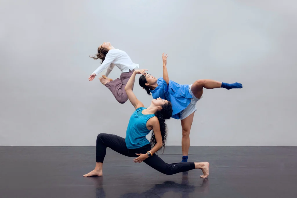 Three dancers pose together. Downstage, a dancer in blue rehearsal clothes lunges deep and arches back. Her upper arm curves toward a dancer balancing on one leg in attitude side. Upstage of them, a dancer caught midair in a C-jump.