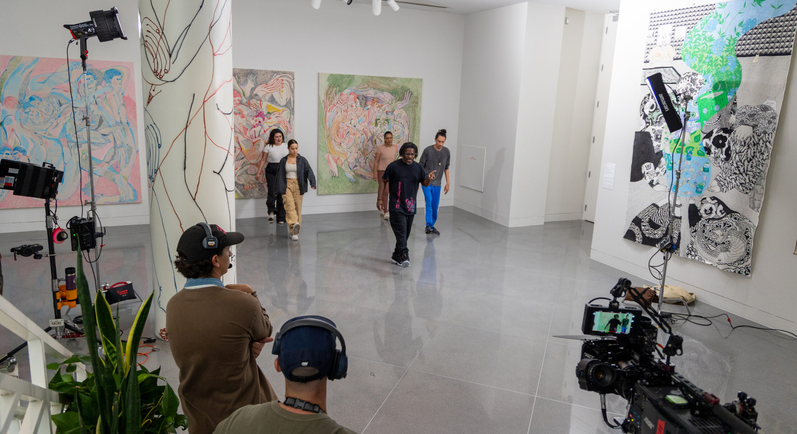 In a vibrant art gallery space, Evans—a dark-skinned man wearing a black t-shirt and black pants—demonstrates a dance move for a group of four teenaged students. In the foreground, the camera crew filming the process is visible.