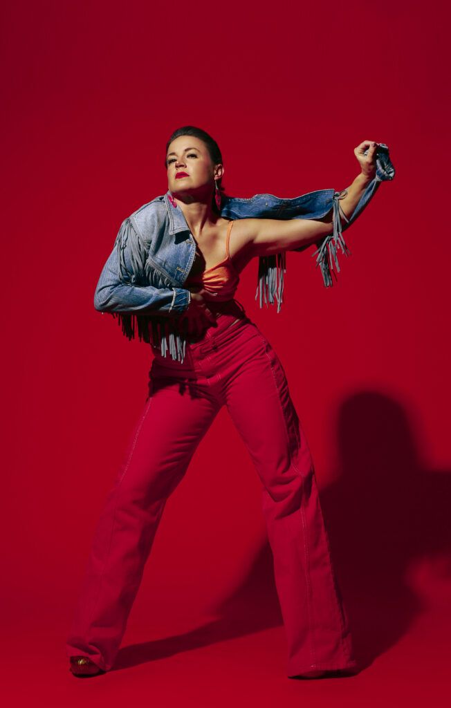 Struxness, wearing bright red pants and a fringed jean jacket, poses against a red background with her left arm extended and her right hand splayed under her ribcage.