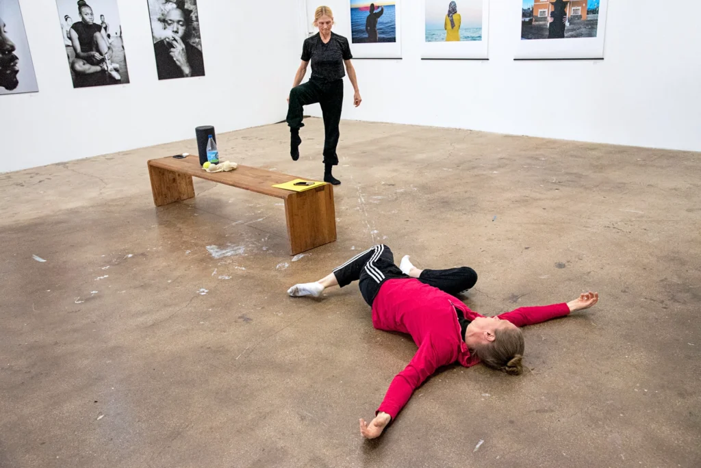 Two dancers warm up in an art gallery. One twists on the floor, the other tests her balance on one leg.
