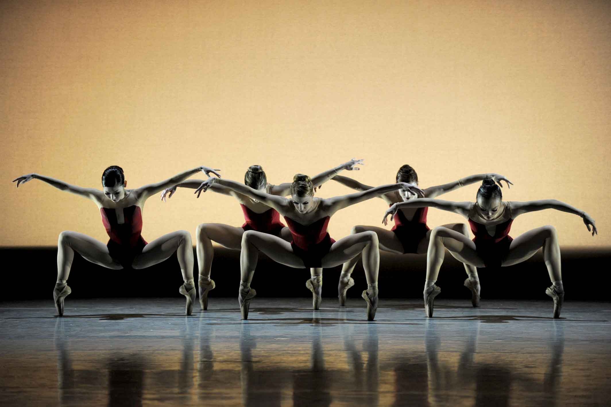 A cluster of five female dancers balance in deep second position pliés on pointe. Their arms extend to the sides like wings, heads bowed to look toward the floor. The backdrop is a faded orange, the stage lighting shadowy.