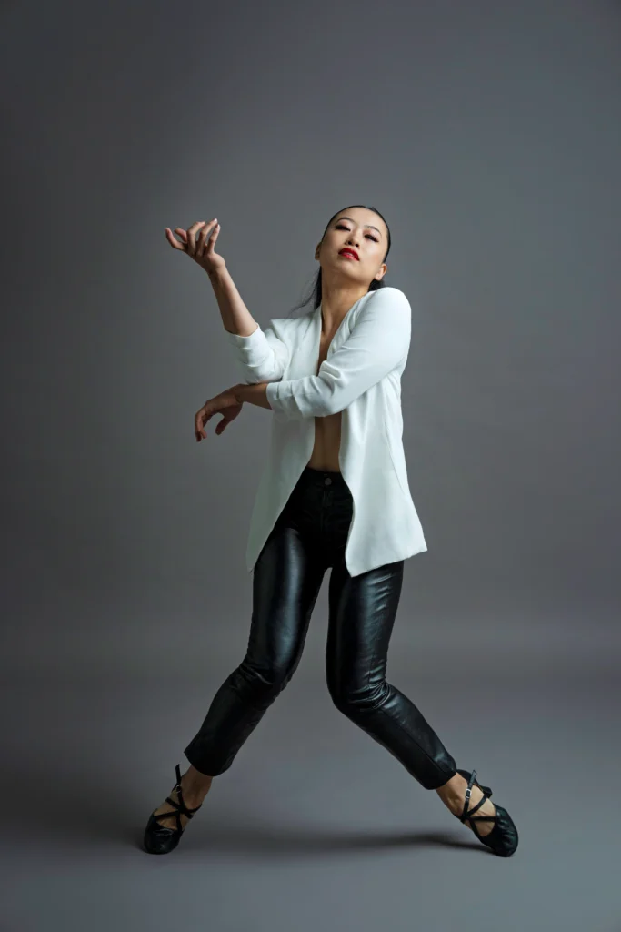 Erina Ueda balances on the tips of her toes in forced arch, knees turning in. She lifts the chin as she regards the camera, arms crossed so one elbow elevates an elegantly raised hand. She wears a white cardigan open over black leather leggings and black heeled jazz shoes.