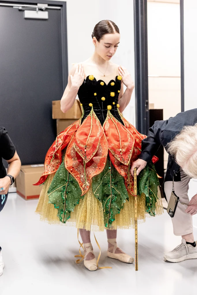 A dancer glances down at the costume she is being fitted in. The bodice is dark, while the calf-length tutu is layered orange, green, and yellow.