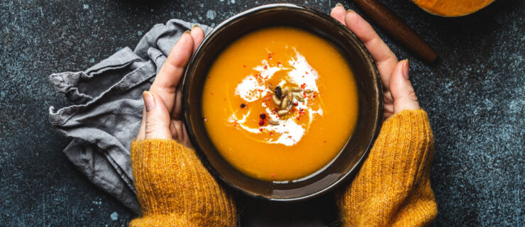 Female hands in yellow knitted sweater holding a bowl with pumpkin cream soup on dark stone background with spoon decorated with cut fresh pumpkin, top view. Autumn cozy dinner concept