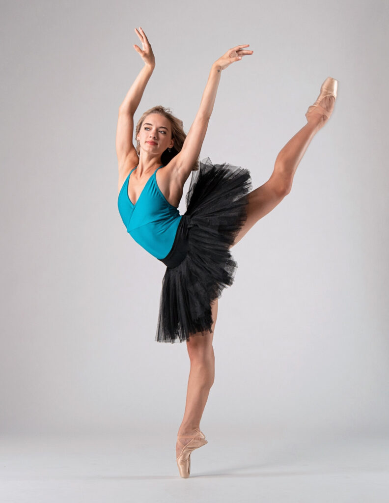 Grace Rookstool balances in back attitude on pointe. Her arms are raised in a soft V similar to Swan Lake. Her blonde hair is loose behind her shoulders. She wears a black practice tutu over a turquoise leotard.